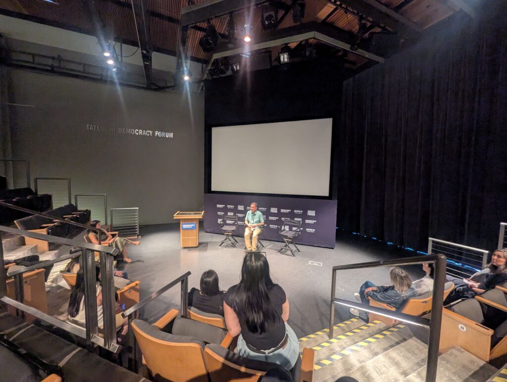 A speaker sits on a stage in a small auditorium, facing an audience. The room is dimly lit with a large screen behind him. The ambiance is intimate and focused.