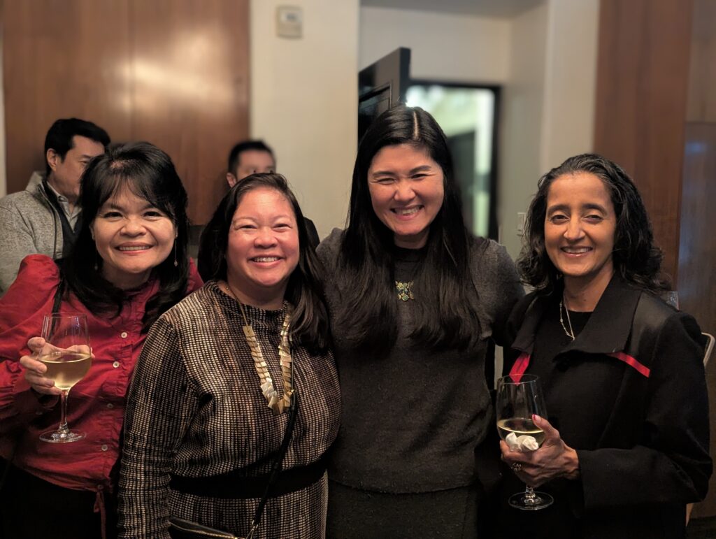 Four women smile together at an indoor social event. From left to right: Chun-Yen Chen in a red blazer holding a wine glass, Stephanie Lomibao in a houndstooth patterned top with statement necklace, Connie Chung-Joe in a dark sweater, and Rochelle Witharana in black attire holding a wine glass. Warm lighting and other attendees are visible in the background.