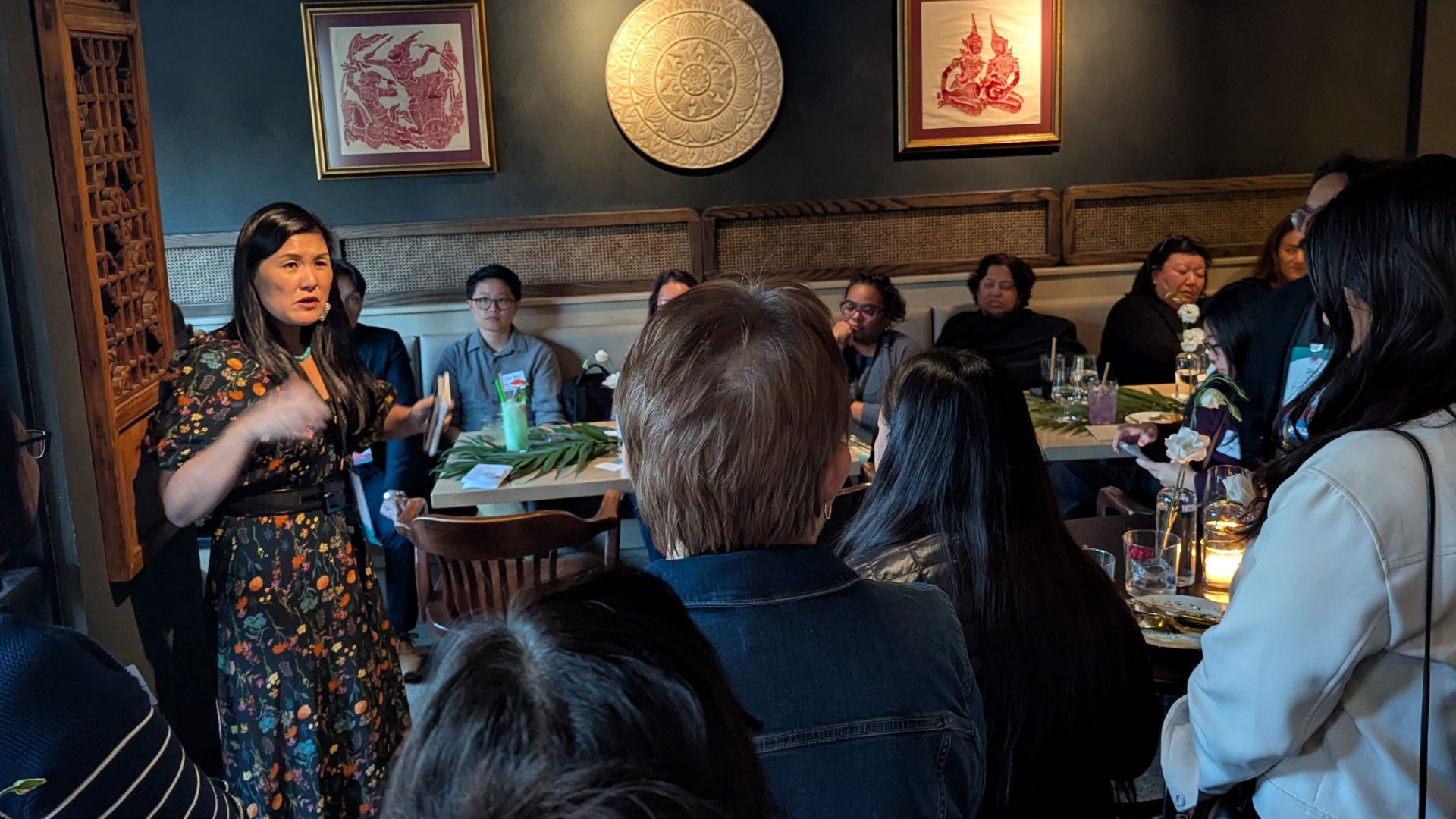 Connie Chung Joe, President and CEO of AAPIP, stands and presents to an audience seated around dining tables in a restaurant setting. She wears a dark floral dress and gestures while speaking. Attendees face her from their seats, with water bottles and table settings visible. The room features decorative wall art including framed Asian motifs and a circular gold ornamental plate. The intimate gathering includes approximately 15-20 people in a professional networking environment.