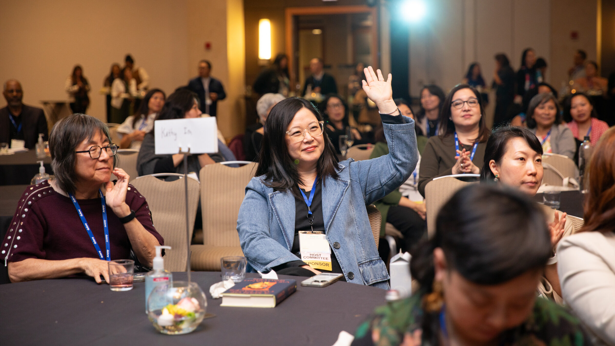 An indoor event photograph taken in a dimly lit hotel ballroom. In the center foreground, a woman wearing glasses and a denim jacket raises her hand, her badge visible and labeled "Host Committee / Sponsor." She is seated at a round table with a dark tablecloth, on which a book, a glass bowl centerpiece, and a hand sanitizer bottle are visible. To her left, another woman in a burgundy top looks on. The room is filled with attendees seated at round tables and standing in the background, most wearing blue lanyards. Stage lighting and an exit sign are visible at the rear of the room.