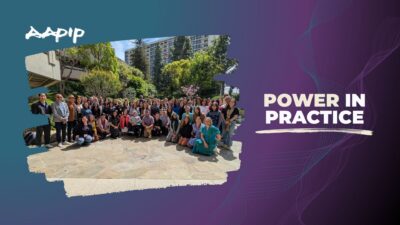 A wide-format 16:9 graphic with a teal-to-purple gradient background and subtle flowing line patterns on the right side. The AAPIP logo appears in white brush-stroke lettering in the upper left. On the left, a group photograph with torn-paper style edges shows approximately 60–70 people gathered on a flagstone courtyard on a sunny day. Participants are arranged in several rows—some standing, some kneeling, and one person seated in a power wheelchair in the center. The group is racially diverse, smiling, and dressed in business casual attire. The background features mature trees, blooming pink cherry blossoms, and a multi-story building above the treeline. On the right side of the graphic, bold white text reads "POWER IN PRACTICE" with a white brush-stroke underline beneath it.