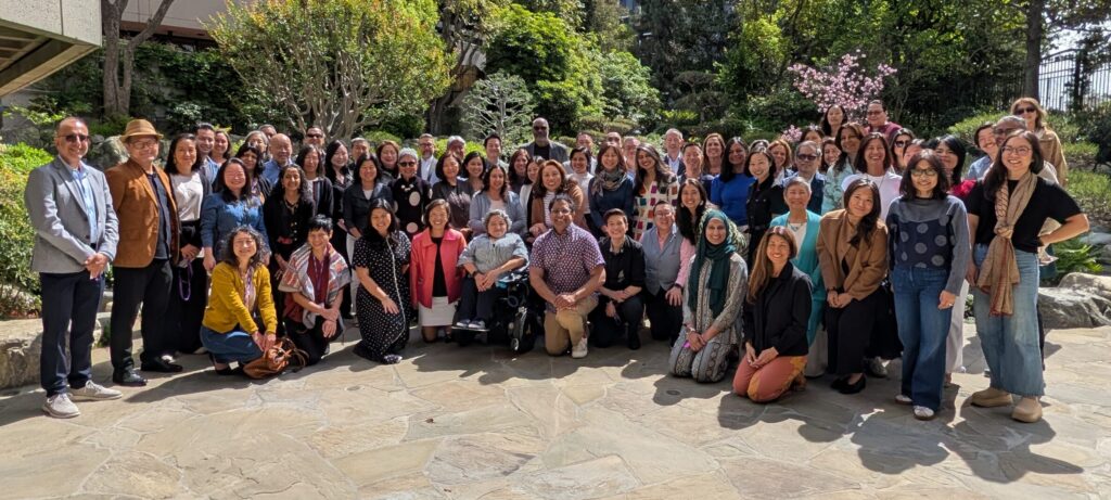 An outdoor group photograph taken on a sunny day. Approximately 60–70 people are gathered on a flagstone courtyard, arranged in several rows—some standing, some kneeling, and one person seated in a power wheelchair in the center foreground. The group is racially diverse and includes people of varying ages. Many are smiling and dressed in business casual or smart casual attire in a range of colors. The background features mature trees, blooming pink cherry blossoms, ornamental shrubs, and a multi-story concrete building visible above the treeline. Natural light is bright and even.