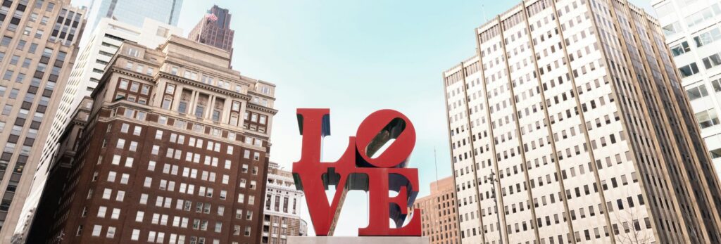 A wide-format photograph taken from a low angle looking up at the iconic LOVE sculpture by Robert Indiana, located in LOVE Park (John F. Kennedy Plaza) in downtown Philadelphia. The large red block-letter sculpture spells "LOVE" in a 2x2 grid with the "O" tilted at an angle. The sculpture is centered in the frame, flanked by mid-century office buildings in beige and white. In the background, a modern glass skyscraper rises against a clear light blue sky. An American flag is visible on a building to the left. The photo is taken on a bright, cloudless day.
