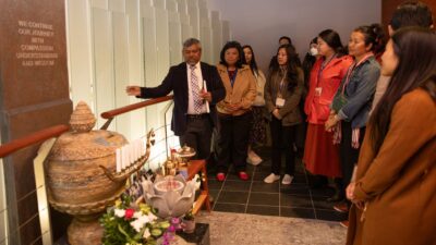 A man in a suit gestures toward a ceremonial altar while speaking to a group of approximately ten people gathered in a memorial or cultural space. A wall inscription reads: "We continue our journey with compassion, understanding and wisdom."