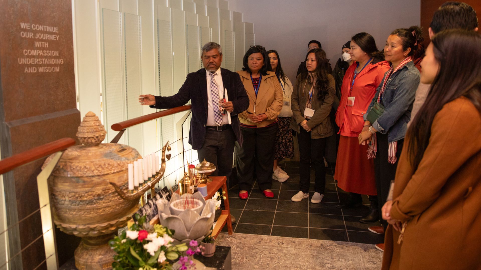 A man in a suit gestures toward a ceremonial altar while speaking to a group of approximately ten people gathered in a memorial or cultural space. A wall inscription reads: "We continue our journey with compassion, understanding and wisdom."