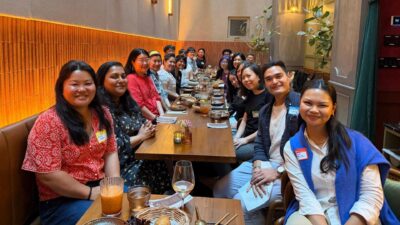 An indoor photograph taken at a long communal dining table in a warmly lit restaurant with amber wall sconces, terracotta tile accents, and a skylight above. Approximately 20 people are seated on both sides of the table, smiling at the camera. Several attendees are wearing "Hello My Name Is" name tags. The table is set with plates, glasses, cocktails, and small floral centerpieces. The group is racially diverse and dressed in smart casual attire.