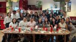 Approximately 30 AAPIP chapter members smile for a group photo around a large oval dining table in a vibrant Asian restaurant, mid-meal, with dishes and red glassware visible.