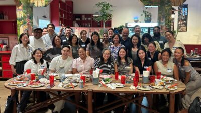 Approximately 30 AAPIP chapter members smile for a group photo around a large oval dining table in a vibrant Asian restaurant, mid-meal, with dishes and red glassware visible.