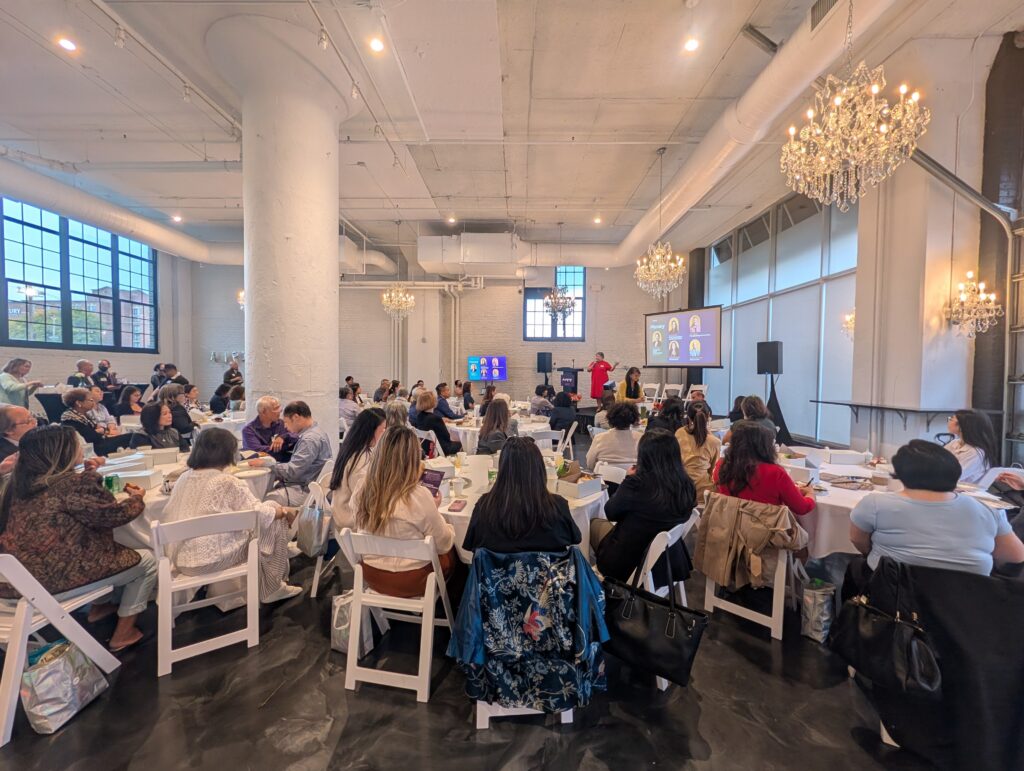 Wide-angle view of an AAPIP Twin Cities event. A full room of approximately 80–100 attendees seated at round tables faces a stage where a speaker stands at a podium. Crystal chandeliers hang from the ceiling of the white-brick venue.