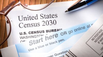 Close-up photograph of a United States Census 2030 mailer from the U.S. Census Bureau, with a pen and reading glasses resting on top. The form reads “Start here OR go online” and “Use a blue or black pen.”